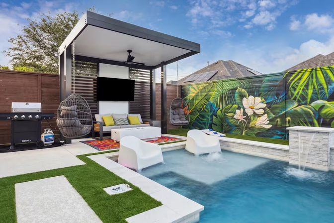 Modern backyard oasis with rectangular pool featuring built-in white lounge seats and waterfall, sleek covered cabana with wall-mounted TV and hanging egg chairs, colorful tropical fence mural and gas grill.