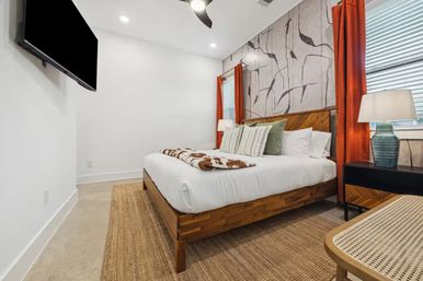 Bright, cozy modern bedroom with a wooden platform bed, white linens, patterned accent wall, orange curtains, woven rug, wall-mounted TV and bedside lamp.