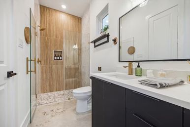 Bright modern residential bathroom with black vanity and white countertop, gold faucet and hardware, glass walk-in shower with vertical wood-look tile accent wall and built-in niche, patterned shower floor, toilet, mirror and small window