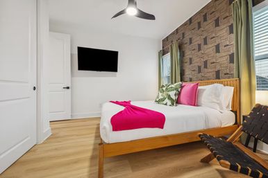 Bright modern bedroom with wooden platform bed, white linens, hot-pink throw and pillows, tropical leaf accent pillow, textured geometric accent wall, wall-mounted TV, hardwood floors, green curtains, woven lounge chair and ceiling fan.