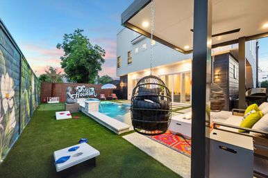 Modern urban backyard at dusk featuring a rectangular plunge pool, black hanging egg chairs, cozy lounge area with fire pit, artificial turf with cornhole boards, and a colorful mural along the fence.