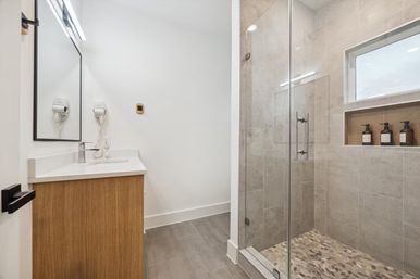Sleek modern bathroom with wood vanity and white countertop, wall-mounted hair dryer, frameless glass walk-in shower with gray tile, pebble shower floor and recessed niche with soap bottles.