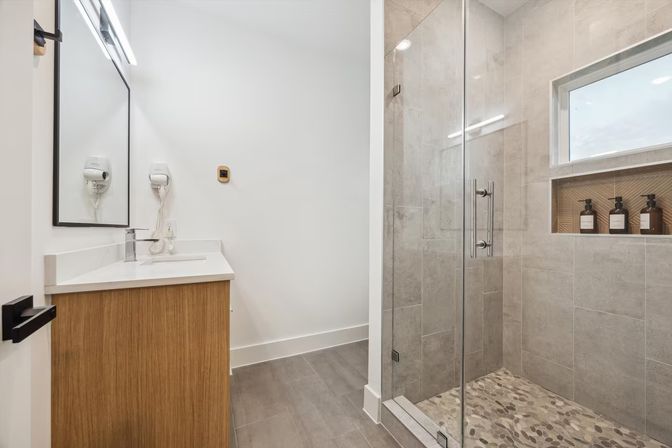 Sleek modern bathroom with wood vanity and white countertop, wall-mounted hair dryer, frameless glass walk-in shower with gray tile, pebble shower floor and recessed niche with soap bottles.