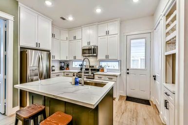 Bright modern kitchen with white shaker cabinets, stainless steel appliances, large island with white quartz countertop, chrome pull-down faucet, wooden stools, and a rear door.