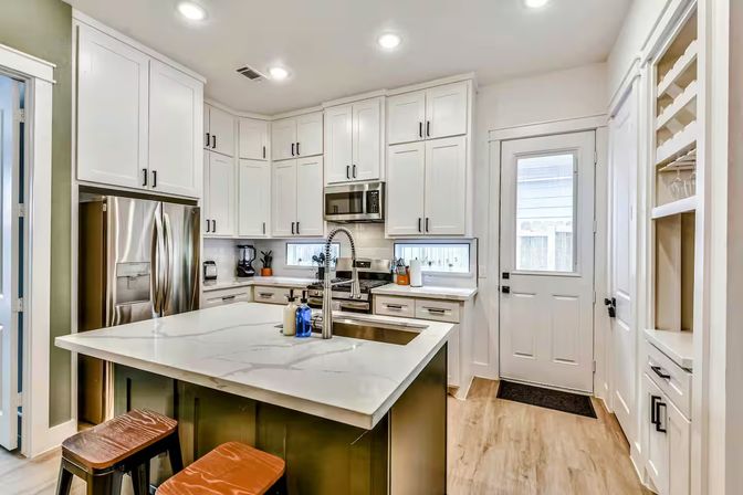 Bright modern kitchen with white shaker cabinets, stainless steel appliances, large island with white quartz countertop, chrome pull-down faucet, wooden stools, and a rear door.