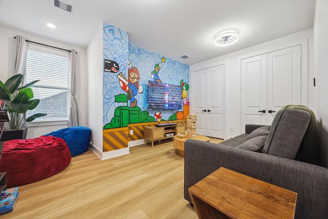 Bright modern family game room featuring a colorful Super Mario wall mural, wall-mounted TV, gray sofa, round wooden coffee table with Jenga tower, red and blue bean bags, hardwood floors and a potted plant