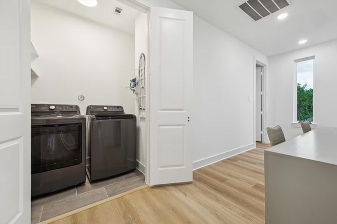 Sleek laundry nook with matching dark gray washer and dryer behind open white closet doors, opening to a bright modern hallway with light wood floors, white walls, and a small desk by a window.