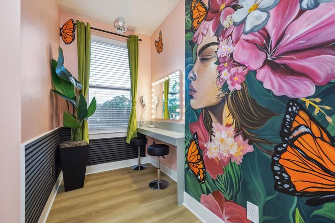 Vibrant beauty room with a large floral mural of a woman's profile and monarch butterflies, lighted makeup vanity and stool, potted plant, green curtains and pink walls.