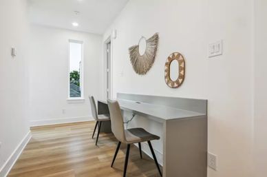 Bright modern hallway home office nook with built-in gray desk, two gray chairs, woven decorative mirrors, tall narrow window and light hardwood floors