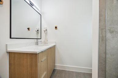Modern minimalist bathroom with wood vanity and white quartz countertop, large black-framed mirror, wall-mounted hair dryer, and gray tiled shower and floor.