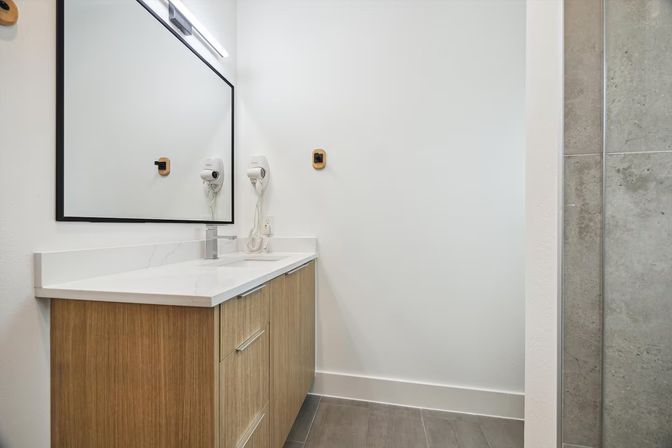 Modern minimalist bathroom with wood vanity and white quartz countertop, large black-framed mirror, wall-mounted hair dryer, and gray tiled shower and floor.