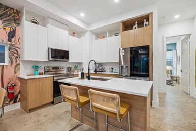 Bright modern open-concept kitchen with white upper cabinets and wood accents, large white island with black pull-down faucet, two cane-and-yellow bar stools, stainless steel fridge and range, and a tropical toucan mural on the accent wall.