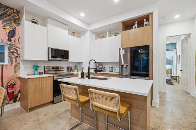 Bright modern open-concept kitchen with white upper cabinets and wood accents, large white island with black pull-down faucet, two cane-and-yellow bar stools, stainless steel fridge and range, and a tropical toucan mural on the accent wall.