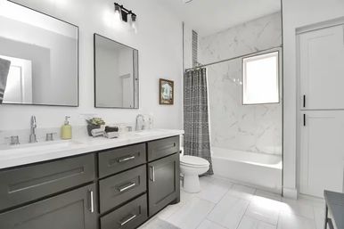 Bright modern bathroom with dark gray double vanity, chrome faucets, marble-tiled tub with patterned gray shower curtain, white tile floor.