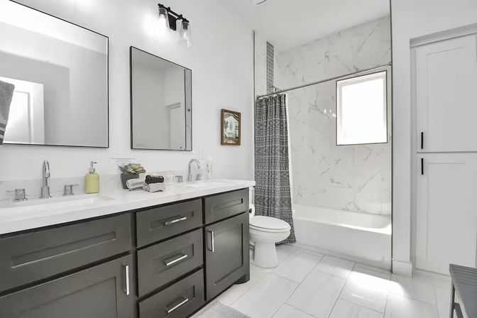 Bright modern bathroom with dark gray double vanity, chrome faucets, marble-tiled tub with patterned gray shower curtain, white tile floor.