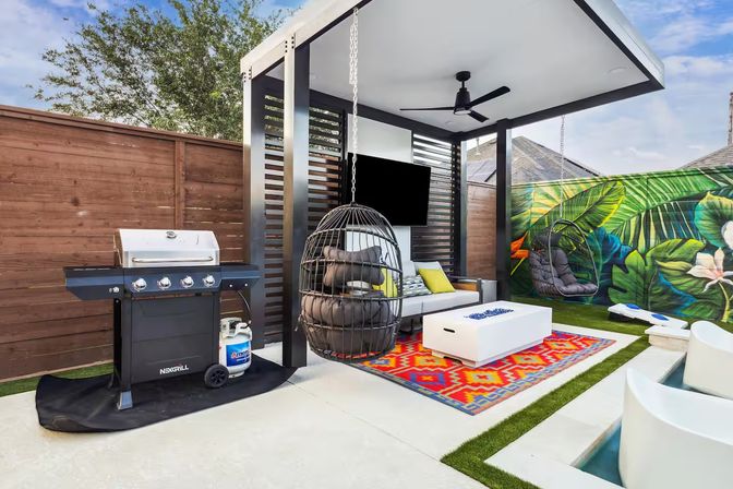 Modern backyard patio with covered pergola, ceiling fan and wall-mounted TV, hanging egg chairs and cushioned sectional on a colorful geometric rug, white coffee table, gas grill with propane tank, tropical leaf mural on the fence and poolside seating.