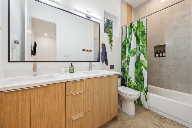 Bright modern bathroom with wood vanity and dual sinks, large framed mirror, tropical green leaf shower curtain over a tiled tub-shower, toilet and small window with a hanging plant