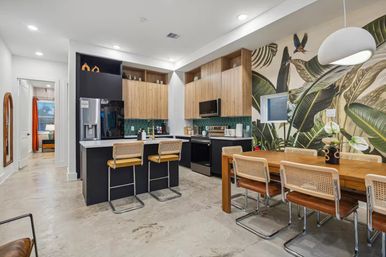 Modern tropical-inspired open-plan kitchen and dining area with a bold botanical wall mural, concrete floors, wood upper cabinets and black lower cabinets, green tile backsplash, marble island with three cane-back bar stools, wooden dining table with cane chairs and a white pendant light.