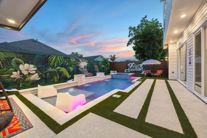 Modern Houston backyard at sunset featuring a glowing rectangular pool with pink LED lounge seats, tropical mural on the privacy fence, concrete pavers with grass strips, and outdoor seating with umbrella.