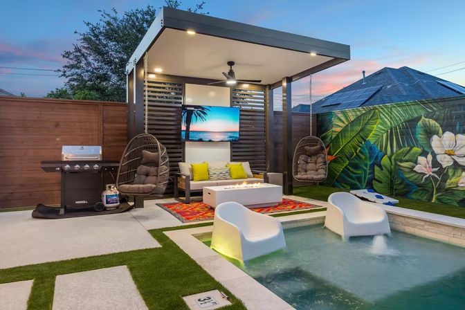 Modern suburban backyard at dusk with a covered pergola, wall-mounted TV, fire-pit coffee table, two hanging egg chairs, gas grill, colorful rug, tropical mural on the fence, and two white in-pool lounge seats in a small plunge pool.