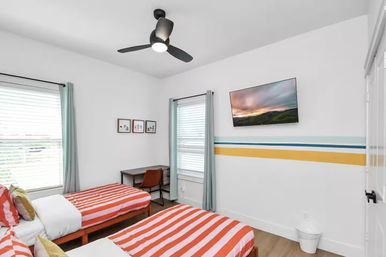 Cheerful modern coastal-style twin bedroom with orange-and-white striped bedding, wall-mounted TV, small desk and chair, black ceiling fan, teal curtains, and blue-yellow accent stripes on white walls