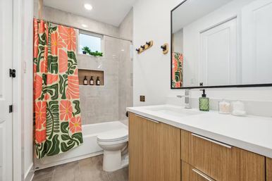 Bright contemporary bathroom with wood vanity and white countertop, large mirror, tub/shower framed by a cheerful coral-and-green floral curtain, neutral tile walls, recessed shower niche with bottles and a small windowsill plant.