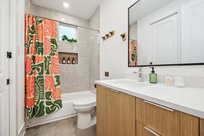 Bright contemporary bathroom with wood vanity and white countertop, large mirror, tub/shower framed by a cheerful coral-and-green floral curtain, neutral tile walls, recessed shower niche with bottles and a small windowsill plant.