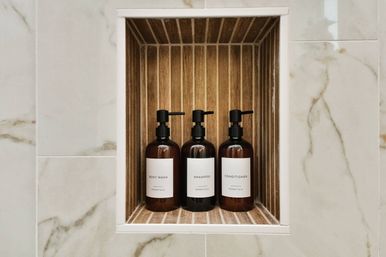 Three amber pump bottles labeled Body Wash, Shampoo, Conditioner arranged in a wood-lined shower niche set into marble-look bathroom tile — modern spa-style bath amenity