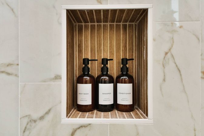 Three amber pump bottles labeled Body Wash, Shampoo, Conditioner arranged in a wood-lined shower niche set into marble-look bathroom tile — modern spa-style bath amenity