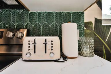 White marble kitchen countertop with a cream four-slice toaster, white paper towel on a wooden holder, textured glass jar and stainless-steel range knobs in front of a green arched leaf-pattern tile backsplash.