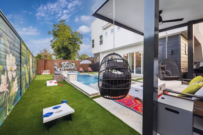 Modern Houston backyard with rectangular plunge pool, artificial turf lawn, colorful mural on wooden fence, hanging wicker egg chairs, cornhole boards and shaded patio seating under a blue sky.