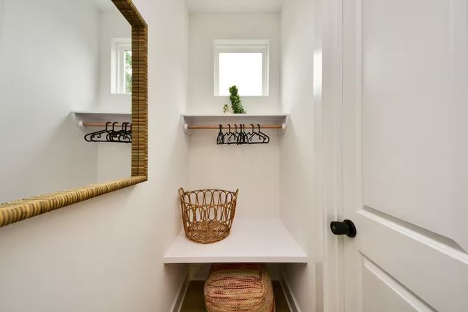 Bright minimalist walk-in closet with natural light, woven rattan laundry basket on a white bench, wall-mounted shelf and wooden rod with black hangers, woven-frame mirror and small window with a potted plant