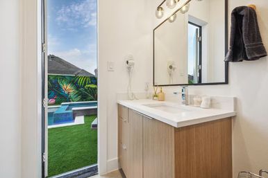 Bright modern bathroom vanity with wood cabinet, white countertop, black-framed mirror and wall-mounted hair dryer; open door reveals backyard pool, artificial turf and colorful tropical mural.