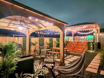 Evening suburban backyard patio with two wooden pergolas draped in string lights, cozy outdoor seating, hammock, potted tropical plants and a raised wooden deck with stairs.
