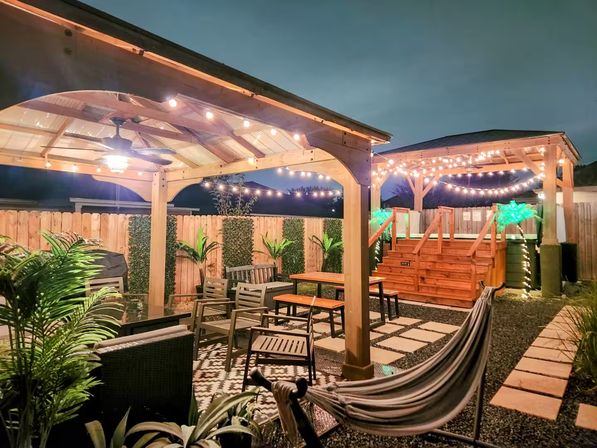 Evening suburban backyard patio with two wooden pergolas draped in string lights, cozy outdoor seating, hammock, potted tropical plants and a raised wooden deck with stairs.
