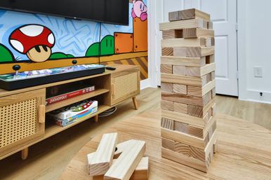 Wooden block tower on a round table with a few removed pieces, in a kids' playroom with a retro arcade controller on a media console, board games on the shelf, and a colorful video-game mural on the wall.