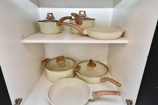 Cream-colored pots and pans with wood-look handles neatly arranged on two shelves inside a white kitchen cabinet
