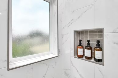 Modern shower with frosted window, white marble-look tiles and a recessed niche with gray mosaic backing holding three amber pump bottles labeled conditioner, shampoo and body wash.