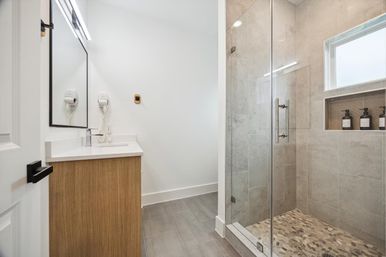 Modern bathroom with wood vanity, white countertop and wall-mounted mirror, sleek glass walk-in shower with pebble-tile floor and recessed niche holding bottles, neutral gray wall tiles and small window.