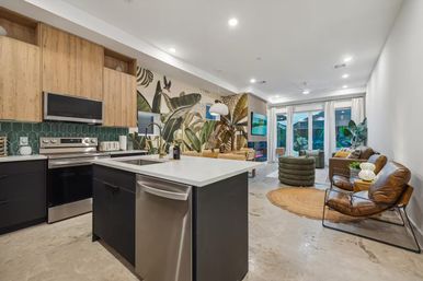 Bright modern open-concept kitchen and living room with white island and sink, stainless steel appliances, wood cabinets, green tile backsplash, bold tropical leaf mural, concrete floors, floor-to-ceiling glass doors, indoor plants and cozy lounge seating.