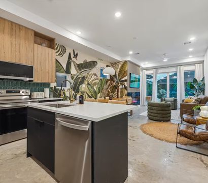 Bright modern open-concept kitchen and living room with white island and sink, stainless steel appliances, wood cabinets, green tile backsplash, bold tropical leaf mural, concrete floors, floor-to-ceiling glass doors, indoor plants and cozy lounge seating.