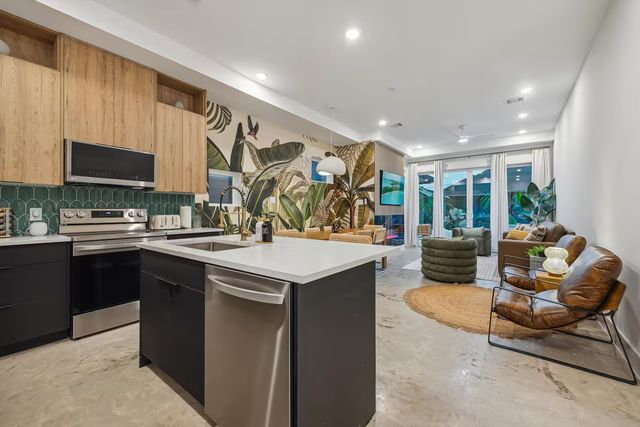 Bright modern open-concept kitchen and living room with white island and sink, stainless steel appliances, wood cabinets, green tile backsplash, bold tropical leaf mural, concrete floors, floor-to-ceiling glass doors, indoor plants and cozy lounge seating.