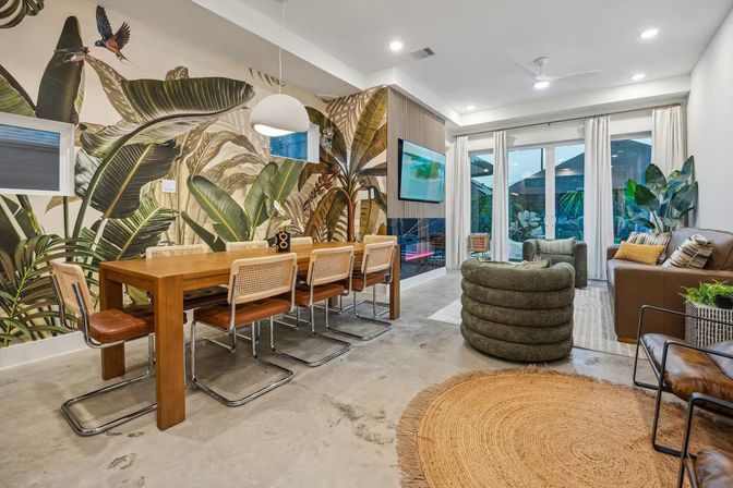 Open-plan modern living and dining room with vibrant tropical leaf mural, solid wood dining table with six cane-back chairs, round jute rug, textured green spiral lounge chair, leather sofa, concrete floor, pendant light, and sliding glass doors to a patio.