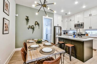 Open-concept modern kitchen and dining area in a residential home with white cabinetry, green accent wall, wood dining table set for six, dark island with stools, stainless steel appliances, hanging sputnik chandelier, and wall plant shelves.