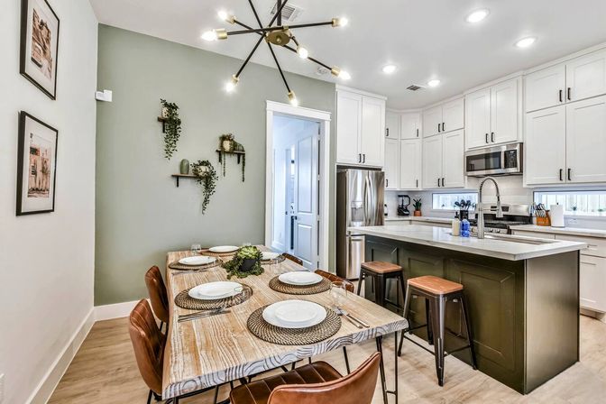 Open-concept modern kitchen and dining area in a residential home with white cabinetry, green accent wall, wood dining table set for six, dark island with stools, stainless steel appliances, hanging sputnik chandelier, and wall plant shelves.