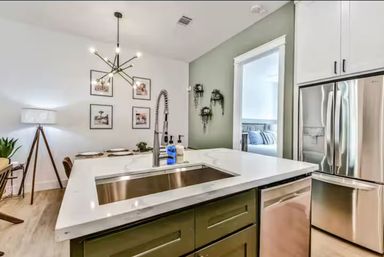 Bright modern open-concept kitchen with white quartz island, stainless farmhouse sink and fridge, olive cabinets, sputnik chandelier, dining nook and green accent wall with wall plants.