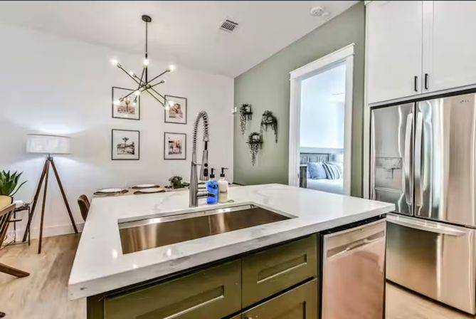 Bright modern open-concept kitchen with white quartz island, stainless farmhouse sink and fridge, olive cabinets, sputnik chandelier, dining nook and green accent wall with wall plants.