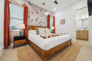 Bright contemporary bedroom with wooden platform bed, abstract botanical accent wall, orange curtains, striped pillows, cowhide throw, woven rug, bedside lamps and visible en-suite bathroom