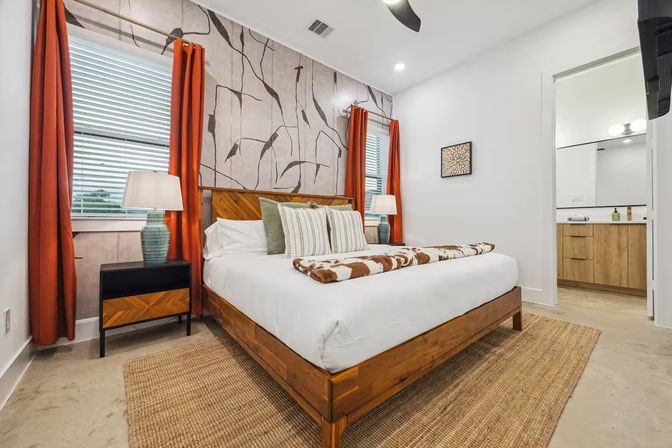 Bright contemporary bedroom with wooden platform bed, abstract botanical accent wall, orange curtains, striped pillows, cowhide throw, woven rug, bedside lamps and visible en-suite bathroom