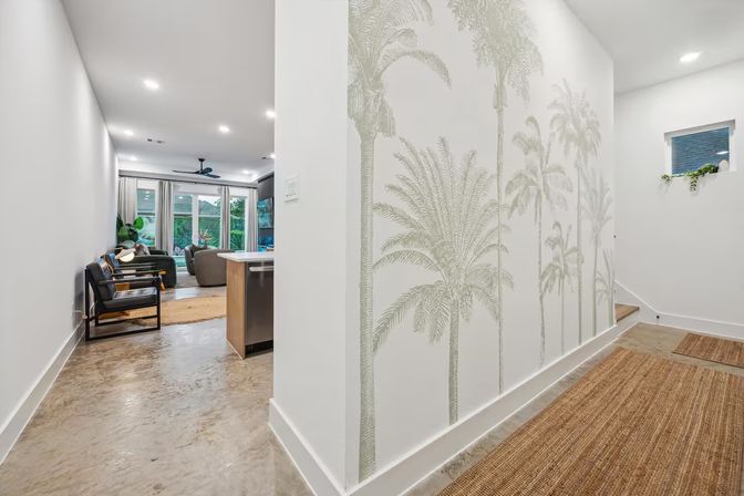 Bright modern hallway with palm-tree wall mural, polished concrete floors and natural fiber runner leading into an open-plan living room with large glass doors, ceiling fan and indoor plants.